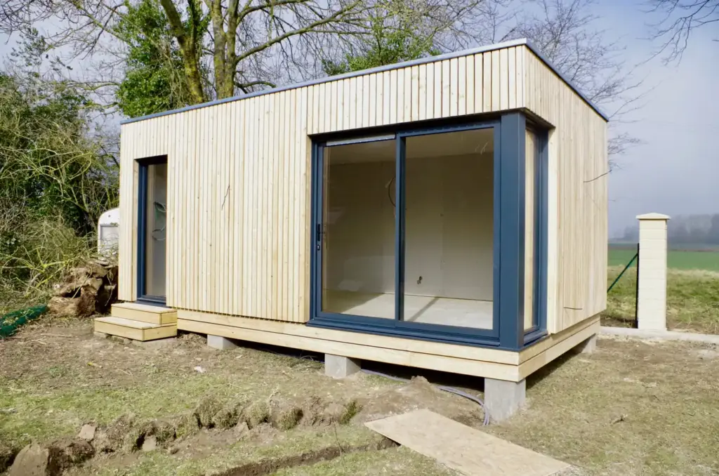 Studio de jardin en bois avec baie vitrée d'angle, posé sur plots dans un jardin