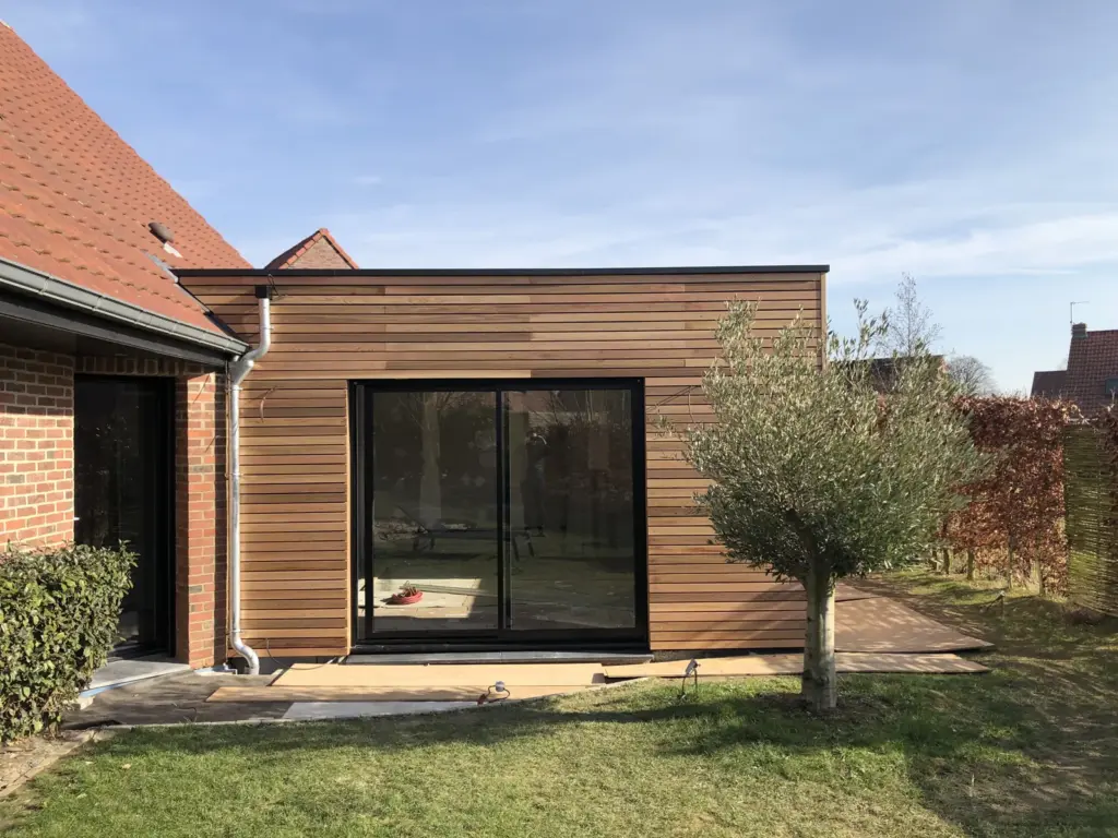 Extension en bois au toit plat accolée à une maison à tuiles rouges, avec un olivier au jardin