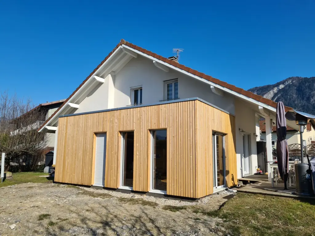 Extension en ossature bois à bardage mélèze vertical, maison à toit de tuiles et montagne au fond
